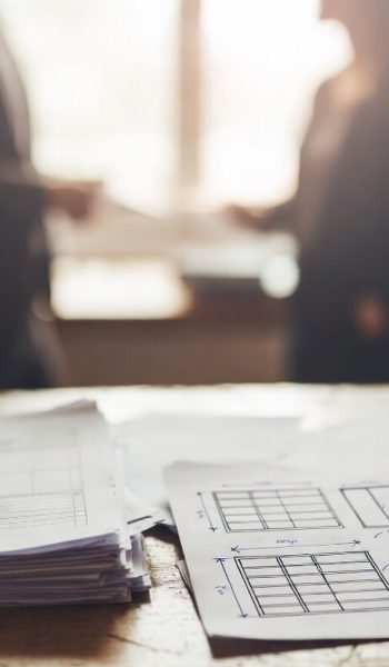 two people having a conversation in the background showing paperwork and sketches on a table in the foreground.